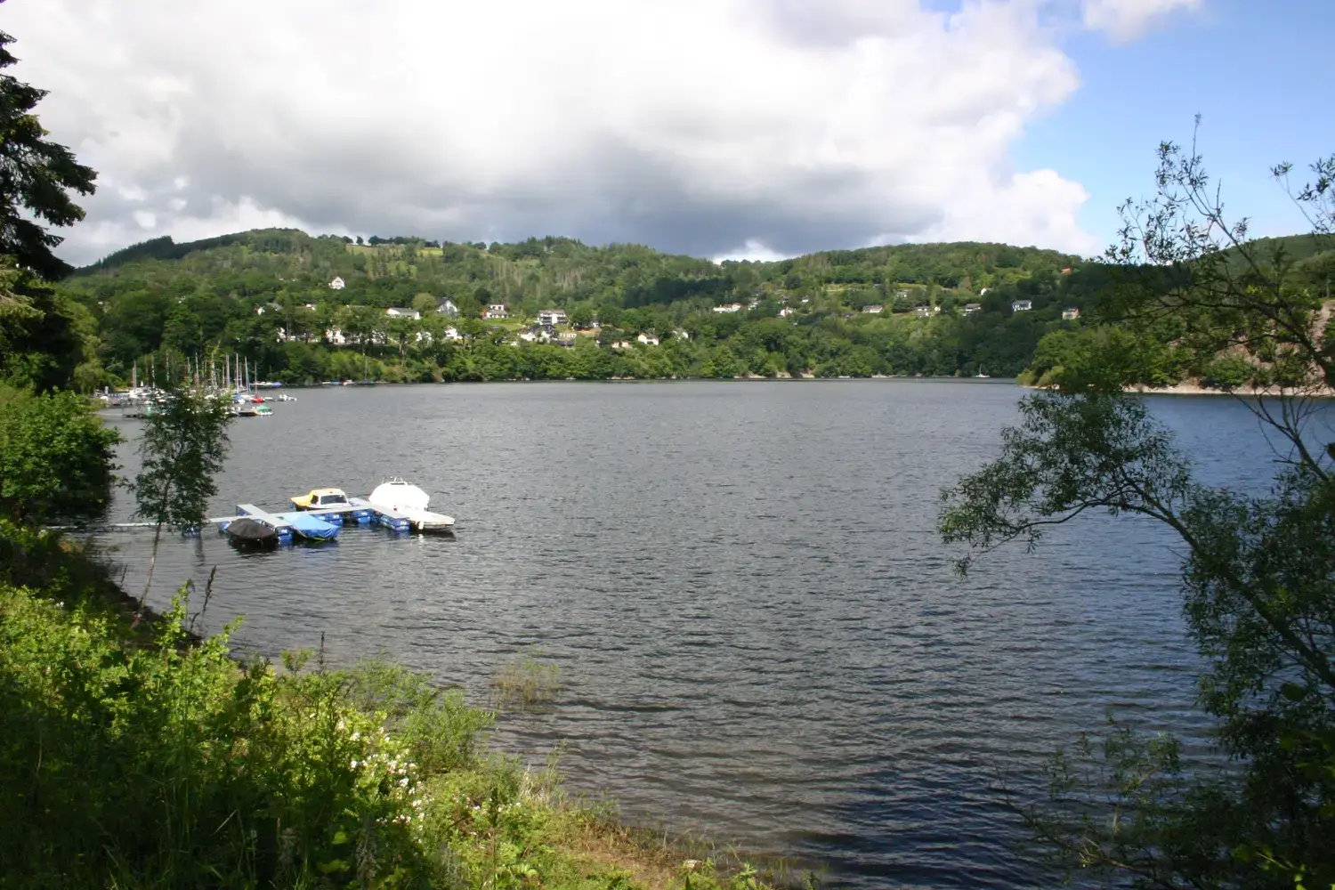 Rursee unter bedecktem Himmel mit Steganlage und Ortslage am gegenüberliegenden Ufer