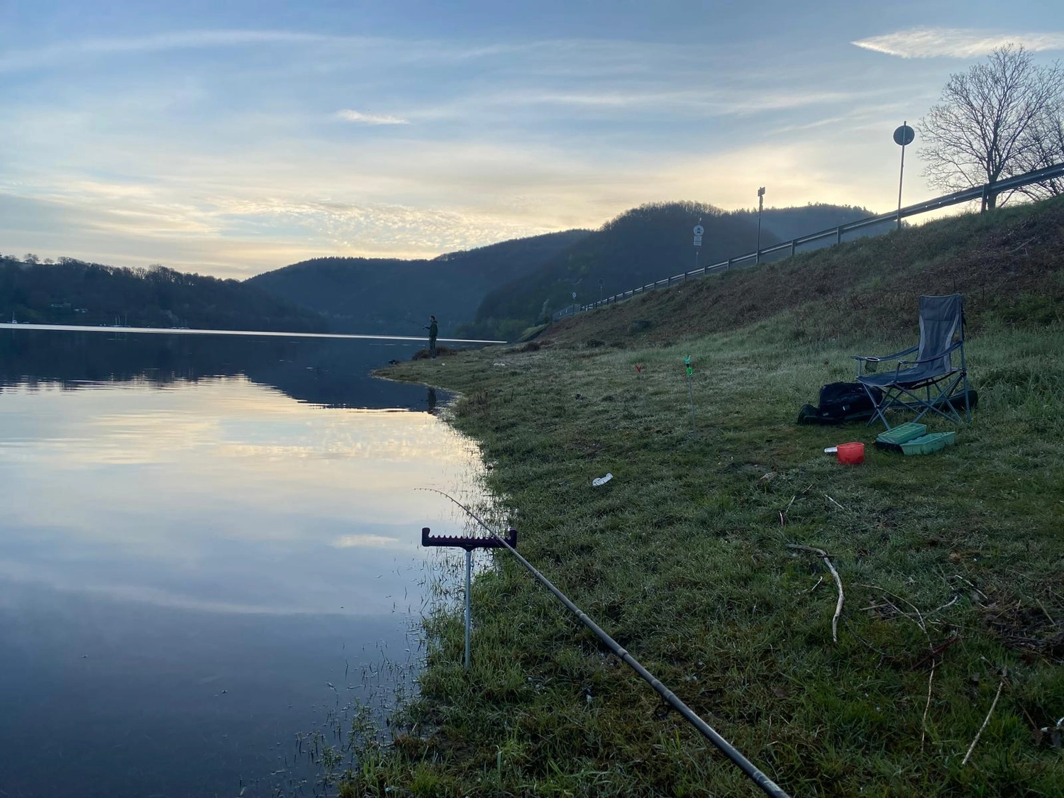 Fischerei-Pächtergemeinschaft Rursee e.V. - Nachhaltige Sportfischerei und Umweltschutz - Eine Angelrute liegt im Morgengrauen an einem ruhigen See auf einem Ständer, daneben befinden sich ein Klappstuhl und Angelausrüstung am grasbewachsenen Ufer.