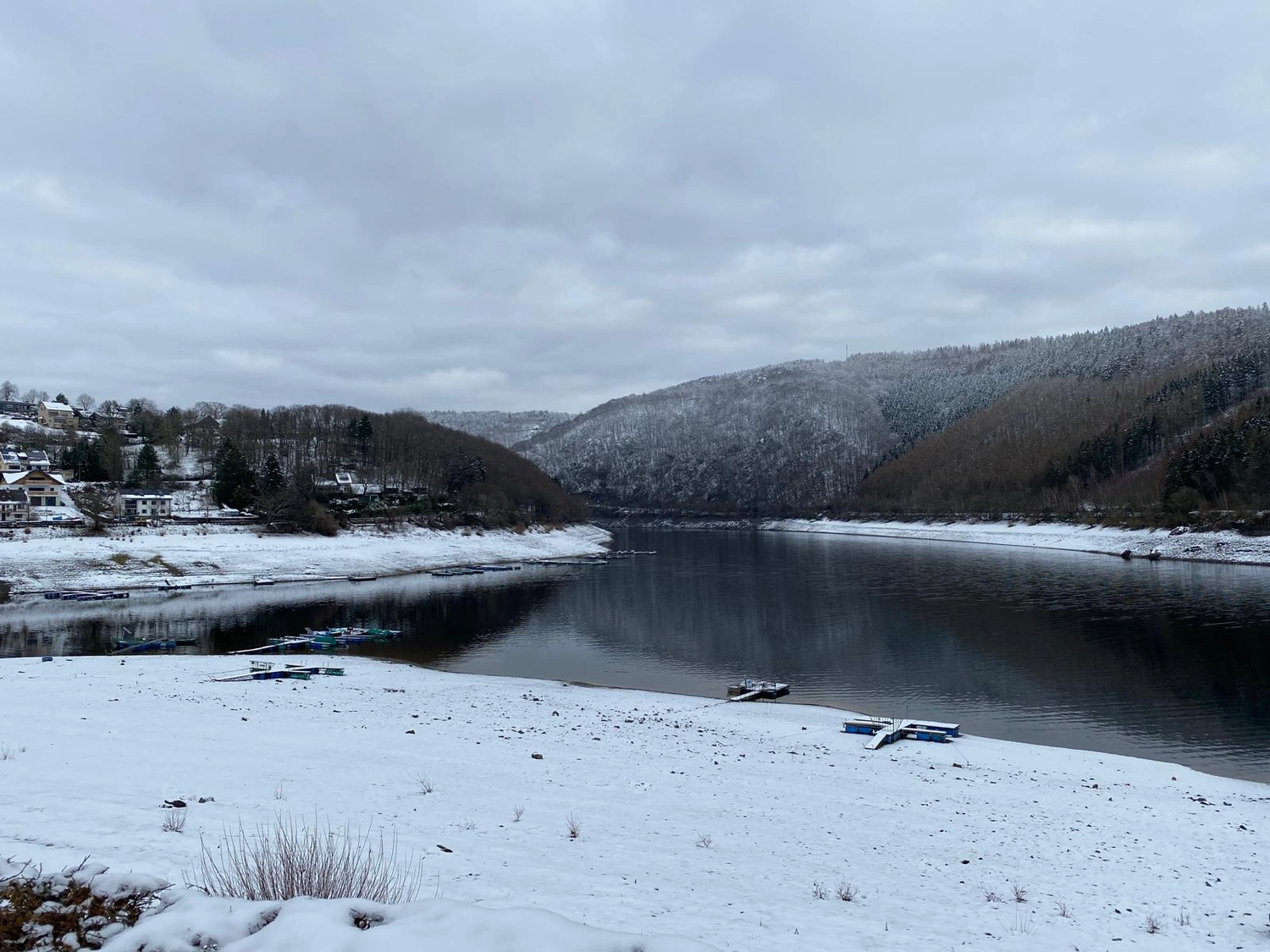 Fischerei-Pächtergemeinschaft Rursee e.V. - Nachhaltige Sportfischerei und Umweltschutz - Eine schneebedeckte Küstenlinie neben einem großen, ruhigen Fluss, der sich unter einem bedeckten Himmel durch bewaldete Hügel schlängelt.