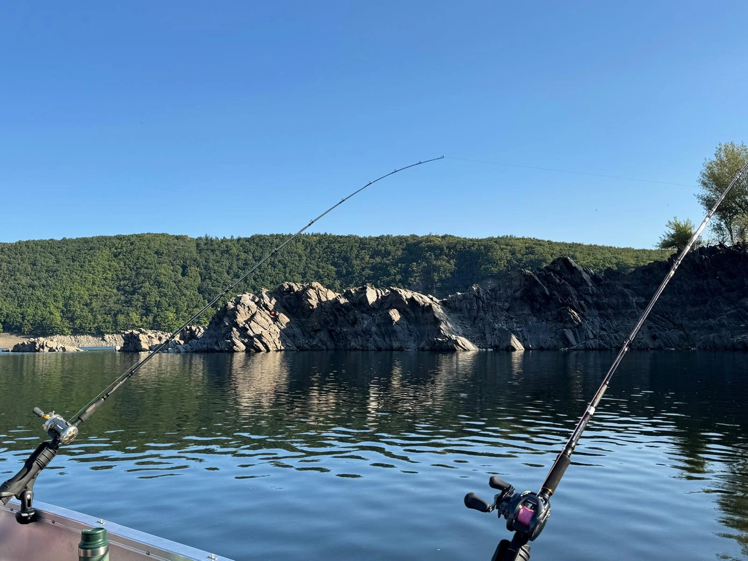 Fischerei-Pächtergemeinschaft Rursee e.V. - Nachhaltige Sportfischerei und Umweltschutz - Zwei Angelruten ragten von einem Boot über das ruhige Wasser, im Hintergrund erhoben sich unter klarem, blauem Himmel felsige Klippen und ein bewaldeter Hügel.