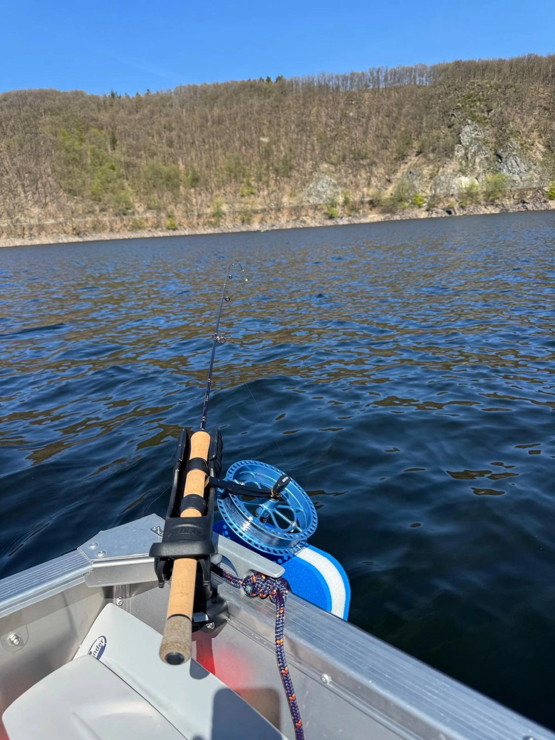 Fischerei-Pächtergemeinschaft Rursee e.V. - Nachhaltige Sportfischerei und Umweltschutz - Eine Angelrute ruht in einer Halterung am Rand eines Bootes und zeigt hinaus auf einen breiten Fluss oder See, im Hintergrund erhebt sich ein bewaldeter Hügel unter klarem blauen Himmel.