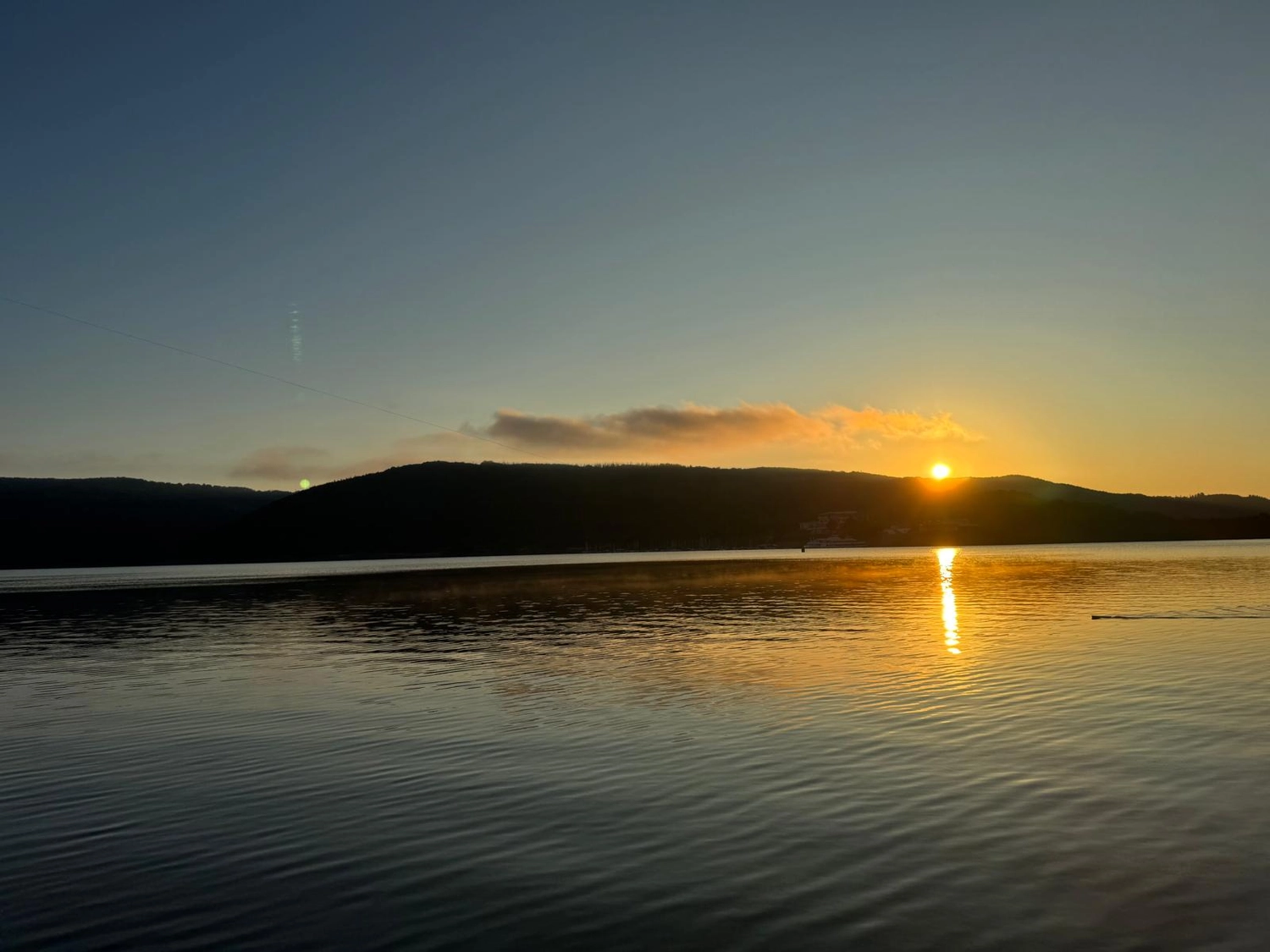 Fischerei-Pächtergemeinschaft Rursee e.V. - Nachhaltige Sportfischerei und Umweltschutz - Bei Sonnenaufgang oder Sonnenuntergang lugt die Sonne über sanfte Hügel und wirft einen leuchtenden goldenen Schatten auf die Wellen eines ruhigen, weiten Sees unter klarem Himmel.