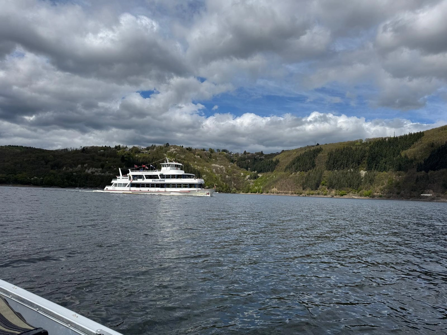 Fischerei-Pächtergemeinschaft Rursee e.V. - Nachhaltige Sportfischerei und Umweltschutz - Ein weißes Passagierboot fährt auf einem breiten Fluss unter bewölktem Himmel; im Hintergrund erheben sich bewaldete Hügel.