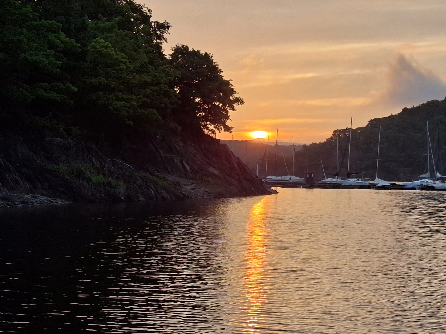 Fischerei-Pächtergemeinschaft Rursee e.V. - Nachhaltige Sportfischerei und Umweltschutz - Ein goldener Sonnenuntergang über einem ruhigen Fluss oder See. Die Sonne versinkt links hinter einem felsigen, bewaldeten Ufer. Das warme Licht erzeugt ein leuchtendes Orange und Gelb, das sich über die Wellen im Vordergrund erstreckt. Im Hintergrund liegen mehrere Segelboote am gegenüberliegenden, fernen Ufer unter einem dunstigen Abendhimmel vor Anker.