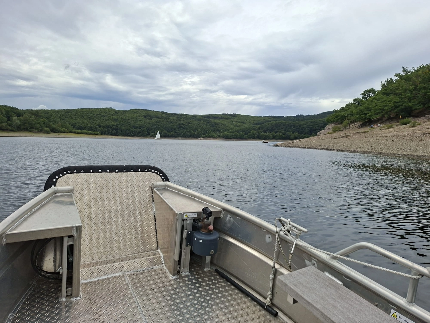 Fischerei-Pächtergemeinschaft Rursee e.V. - Nachhaltige Sportfischerei und Umweltschutz - Ein kleines Aluminiumboot gleitet auf einem ruhigen See unter bewölktem Himmel, in der Ferne ist in der Nähe bewaldeter Ufer ein Segelboot zu sehen.