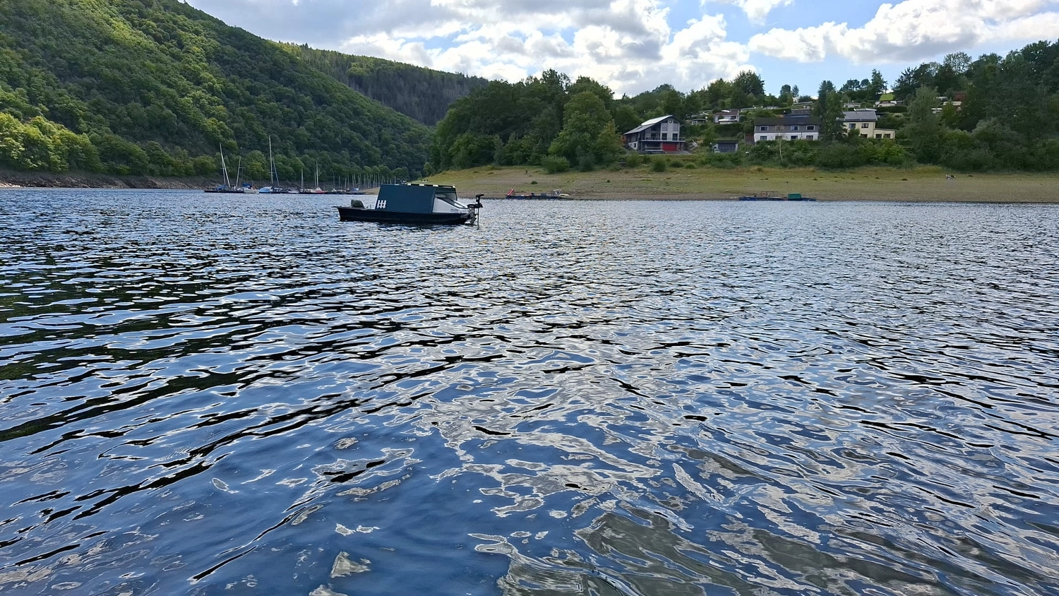 Fischerei-Pächtergemeinschaft Rursee e.V. - Nachhaltige Sportfischerei und Umweltschutz - Ein kleines schwarzes Motorboot treibt auf einem großen, welligen Gewässer; im Hintergrund erheben sich ein bewaldeter Hügel und Häuser am Ufer.