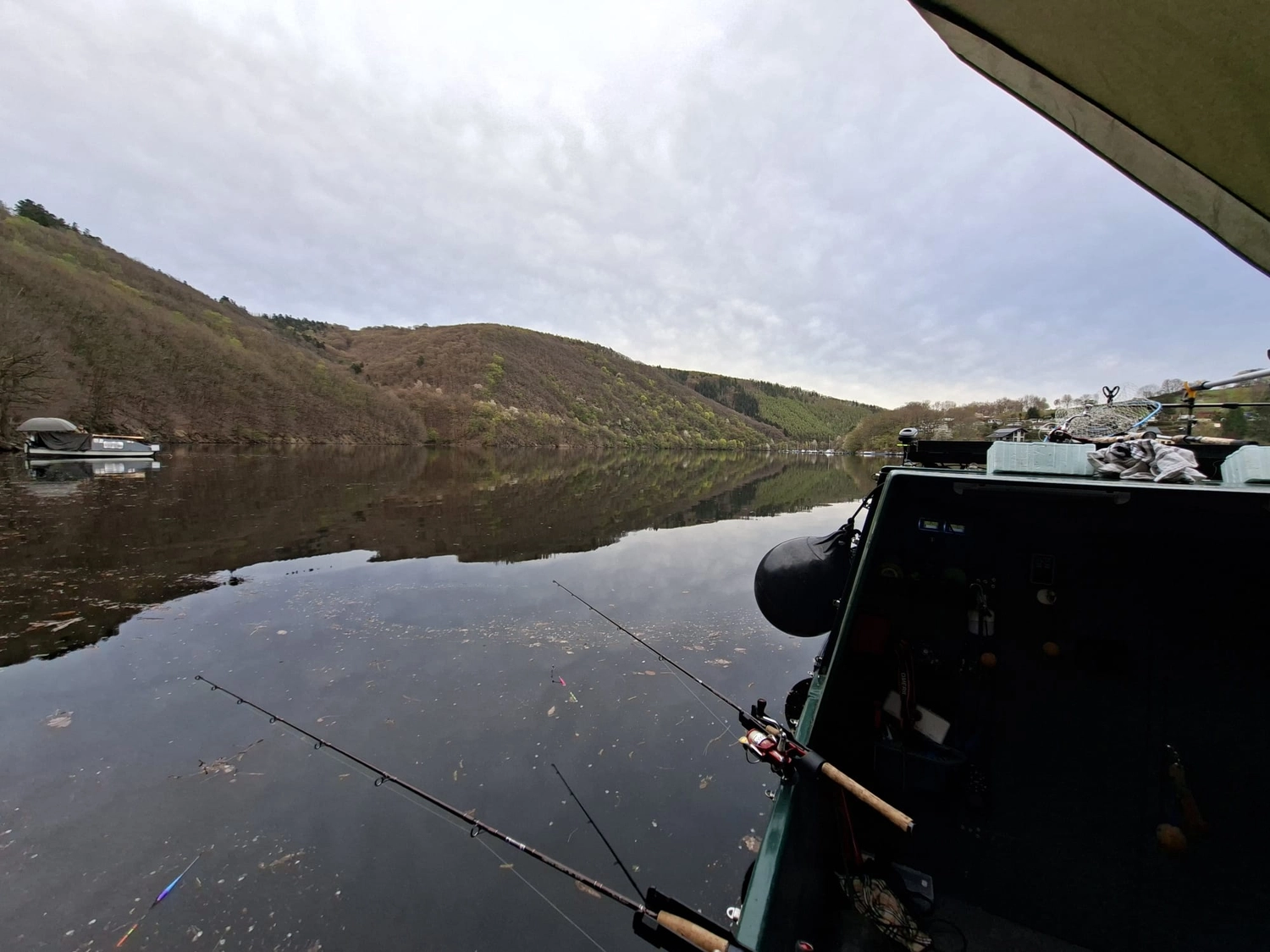 Fischerei-Pächtergemeinschaft Rursee e.V. - Nachhaltige Sportfischerei und Umweltschutz - Blick von einem kleinen Boot auf einem ruhigen, spiegelglatten Fluss, umgeben von bewaldeten Hügeln unter bedecktem Himmel, mit ins Wasser geworfenen Angelruten.